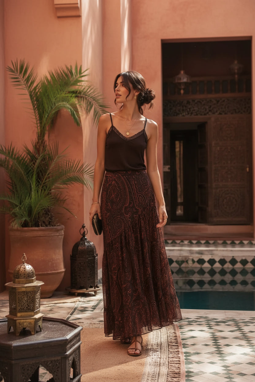 A woman stands in a Moroccan-style courtyard wearing a dark brown camisole and a matching long paisley-print maxi skirt. The setting features terracotta walls, potted plants, and a tiled floor.
