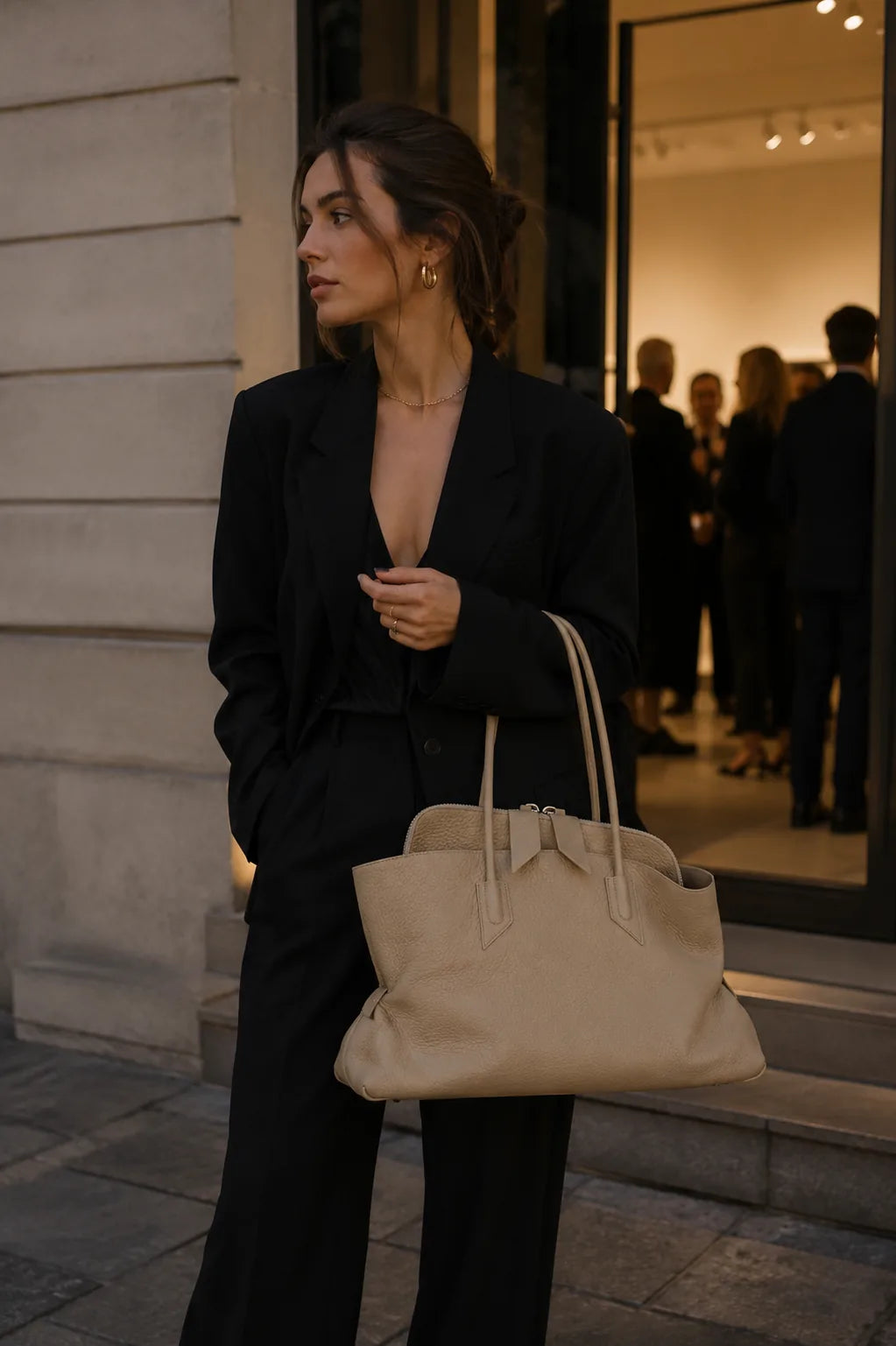 A woman in a black blazer and trousers carries a large beige leather tote bag while standing outside a boutique with a glass entrance and blurred figures inside.